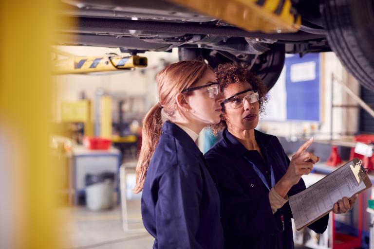 Two mechanics wearing safety goggles looking at the underside of a car that is on a vehicle lift