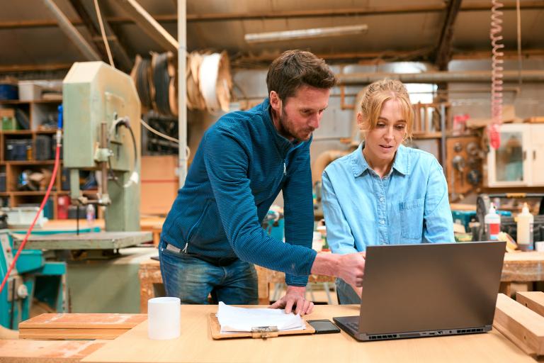 A man and a woman look at a computer at a bench in a workshop