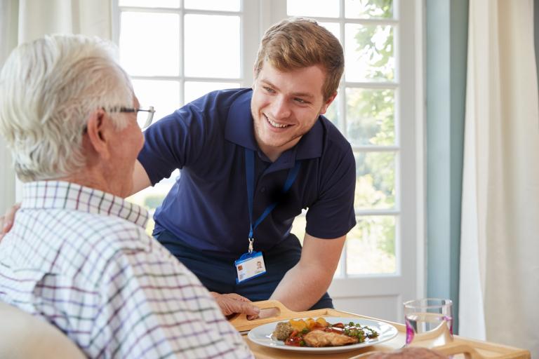 A friendly carer wearing a badge on a lanyard and serving a meal to an older man